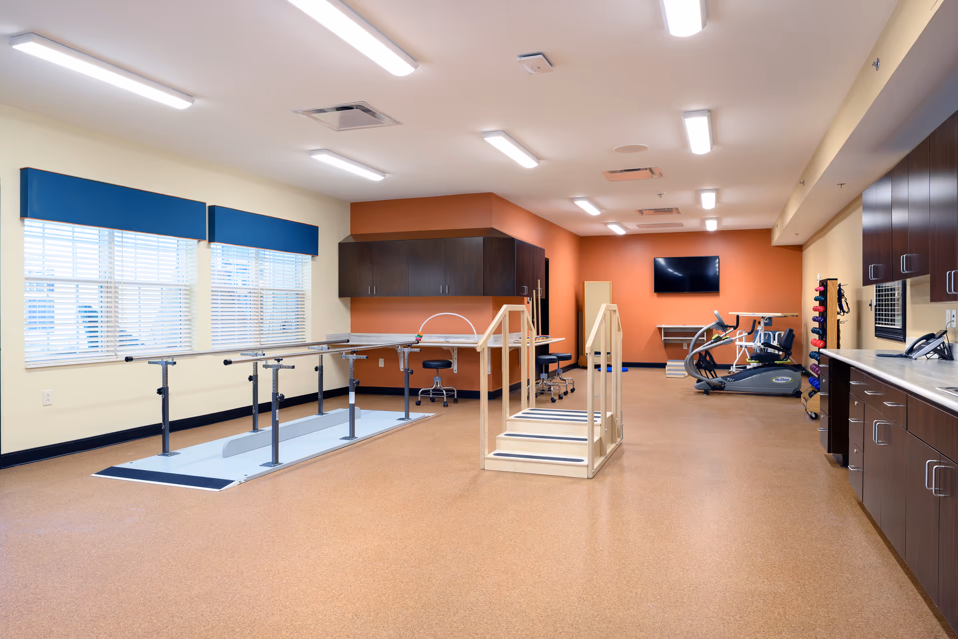 A spacious rehabilitation or physical therapy room with parallel bars for walking exercises, a small set of stairs with handrails, exercise equipment including a treadmill, and cabinets along the walls. The room has bright overhead lighting, windows with blue valances, and a wall-mounted TV.