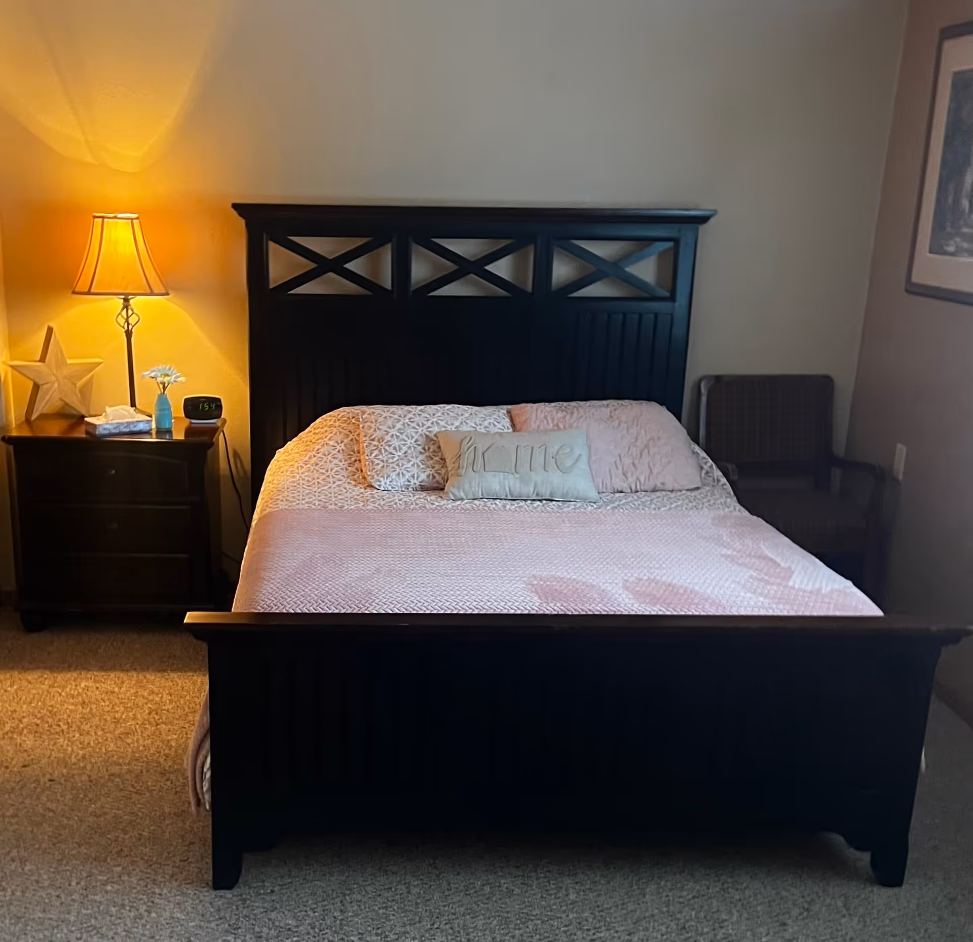 A bedroom with a dark wooden bed frame and headboard, a bed made with light pink bedding and pillows including one with the word 'Home'. To the left of the bed is a nightstand with a lit table lamp, a star decoration, a small vase with a flower, a tissue box, and a digital clock showing 1:54. To the right of the bed is a dark cushioned chair and a framed picture on the wall.