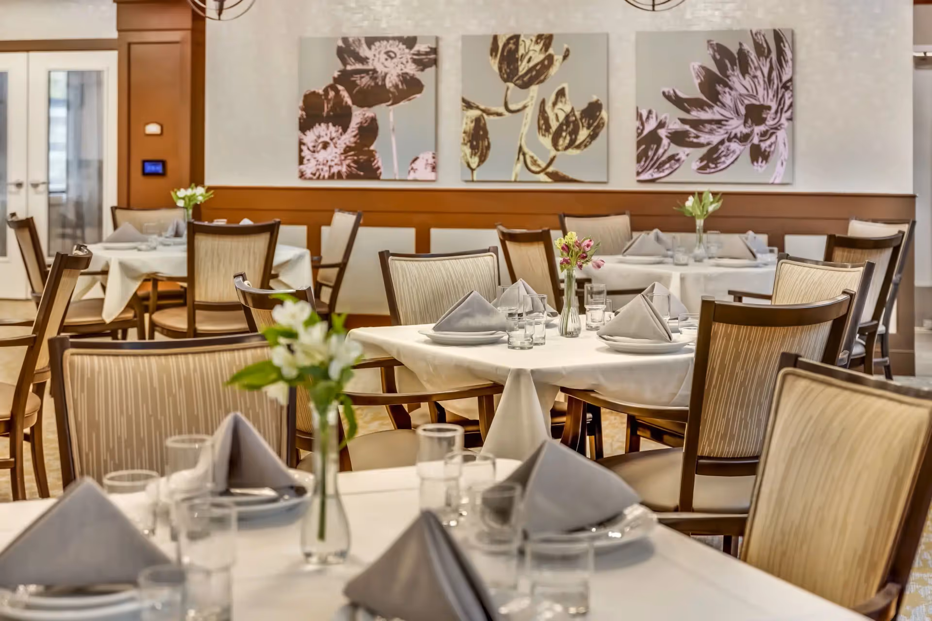 Elegant dining room with multiple tables set with white tablecloths, folded napkins, glassware, and small floral centerpieces.