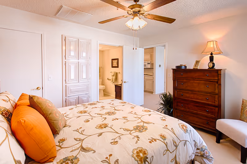 A cozy bedroom in a senior living facility featuring a bed with floral bedding and orange and green pillows. The room has a wooden dresser with a lamp and decorative items on top, a ceiling fan with lights, and an armchair. Two open doors reveal a bathroom with a toilet and towel rack, and a kitchen area with white cabinetry and appliances.