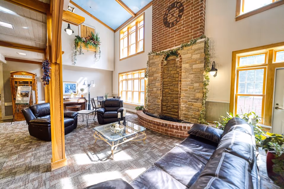 A bright and spacious living room area with large windows allowing natural light to fill the space. The room features a stone and brick fireplace decorated with greenery, a glass coffee table with decorative items, black leather sofas and armchairs, a piano, and various plants. The ceiling is high with wooden beams and a sky-blue painted section.