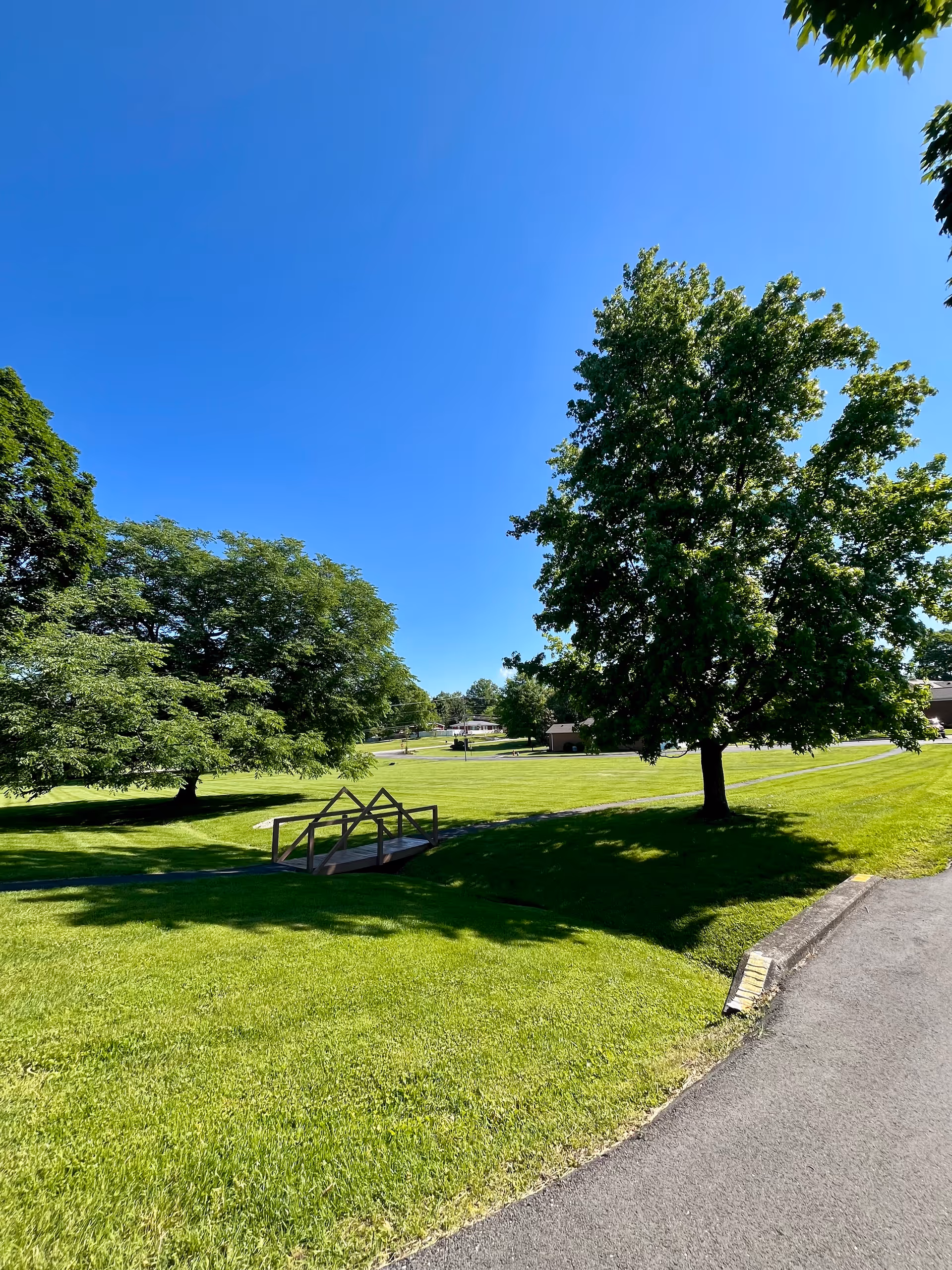 A bright, sunny outdoor scene at Village At Wesley Manor featuring a well-maintained grassy area with several large green trees, a small wooden footbridge over a shallow ditch, and a paved pathway curving along the right side. The sky is clear and blue.