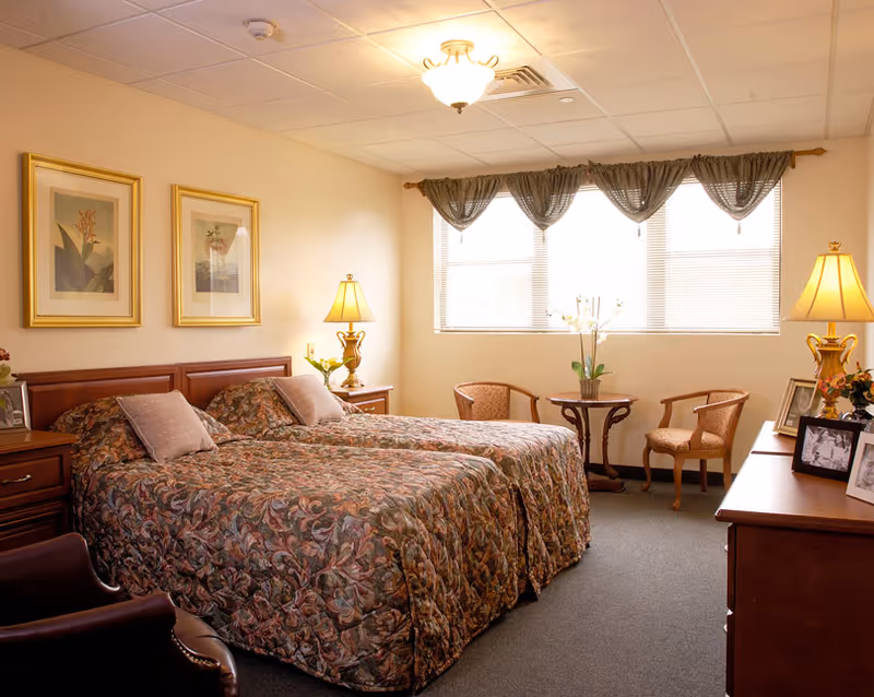 A warmly lit bedroom with two twin beds covered in matching floral bedspreads. Each bed has a pillow and is flanked by wooden nightstands with lamps. The room features a window with sheer curtains, two framed botanical prints on the wall, a small round table with two chairs near the window, and a wooden dresser with framed photos and a lamp.