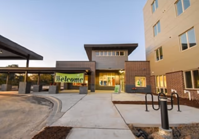 Entrance of a modern assisted living and memory care facility with a covered drop-off area, a large 'Welcome' banner, and a multi-story building with brick and beige siding under a clear sky.