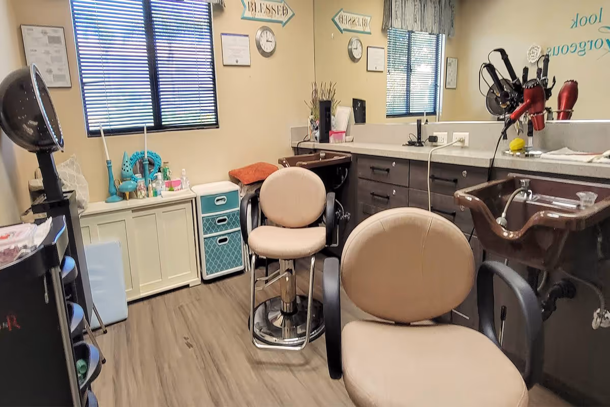 Interior view of a hair salon area within a senior living facility, featuring two beige salon chairs in front of a counter with sinks and hair dryers. The room has wood flooring, a window with blinds, and various hair care items and decorations on the counter and shelves.