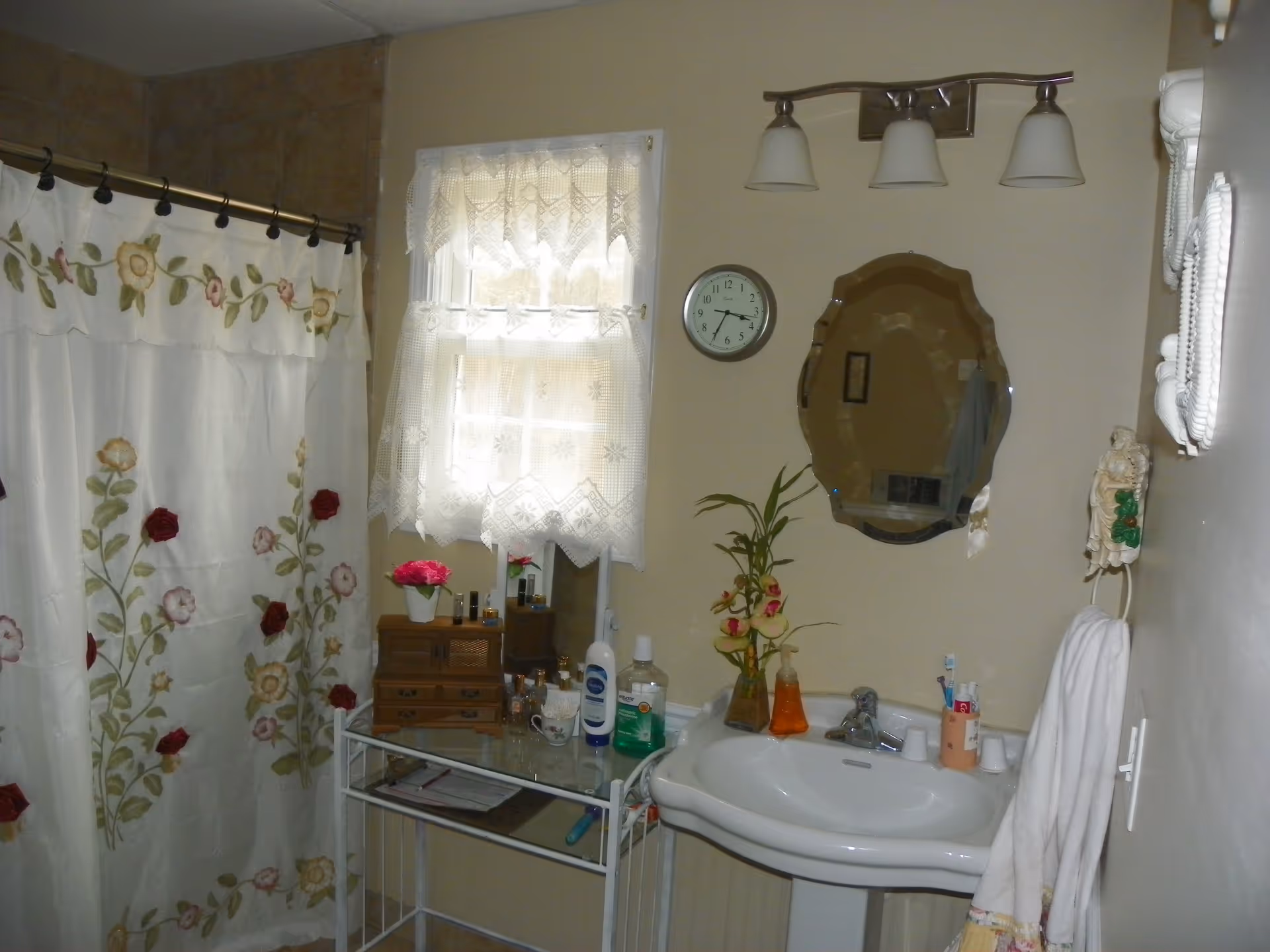 A bathroom with a floral shower curtain, a window with lace curtains, a wall clock, a decorative mirror, a pedestal sink with various toiletries, a towel hanging on a ring, and a small table holding more personal care items and a flower vase.