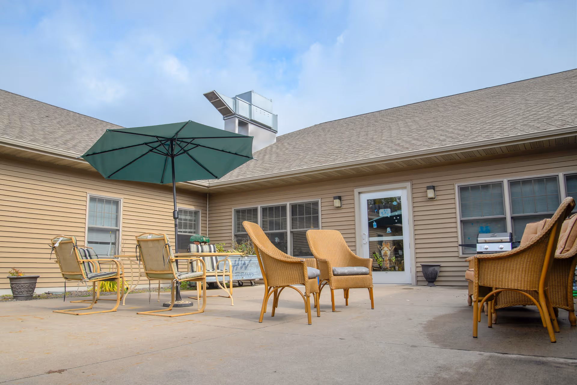 Outdoor patio area at VitaCare Living of Proctor with several chairs and a table under a large green umbrella, surrounded by beige building walls with windows and a door.