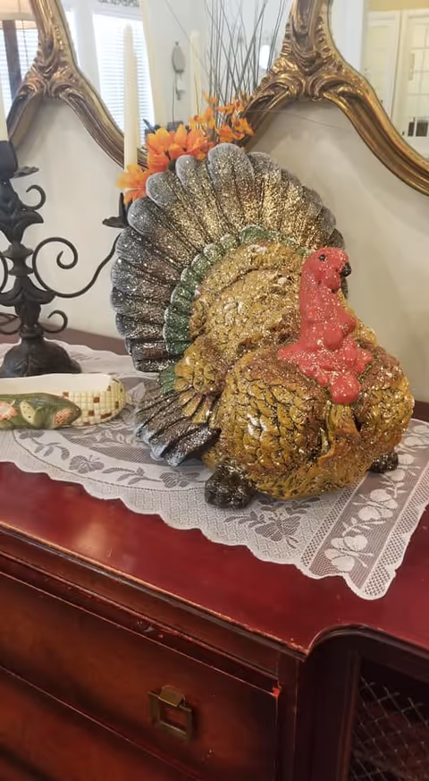 Decorative glittery turkey figurine on a lace-covered wooden sideboard beneath a mirror and next to a candlestick.