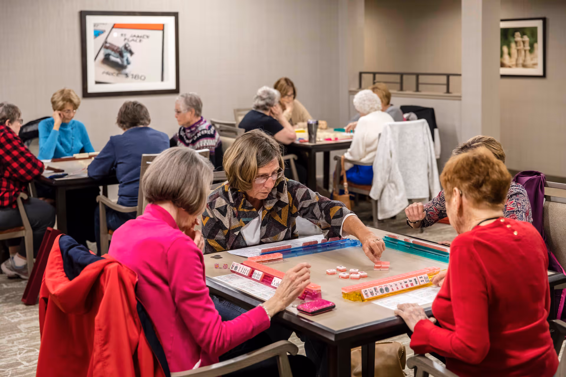 A group of elderly women sitting around tables in a well-lit room playing a tile-based game, likely Mahjong. The room has neutral-colored walls with framed artwork and carpeted floors. The women appear engaged and focused on the game.