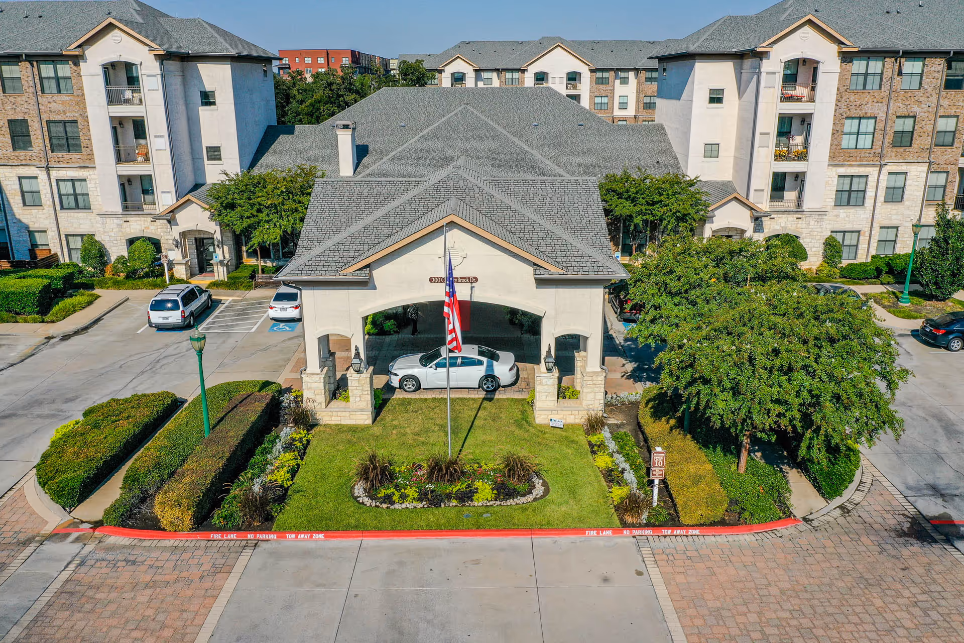 Aerial view of the entrance to a senior living facility named Conservatory At Keller Town Center, featuring a covered drop-off area with a white car parked underneath, an American flag on a flagpole in front, neatly trimmed bushes, trees, and multi-story residential buildings in the background.