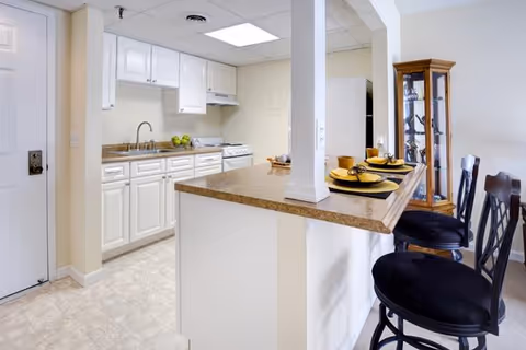 Bright kitchen area with white cabinets, a countertop with a sink, stove, and a breakfast bar with three black cushioned stools. The breakfast bar is set with yellow plates and cups. A glass display cabinet is visible in the background.