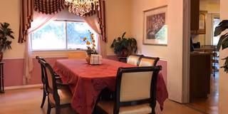 Dining room with a rectangular table covered with a red tablecloth surrounded by six chairs. A chandelier hangs above the table. There is a window with curtains on the left wall, a framed picture on the right wall, and plants placed around the room. The room has light-colored walls and wooden flooring.