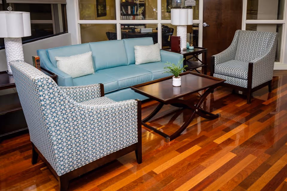 A cozy seating area with a light blue sofa, two patterned armchairs, a wooden coffee table with a small potted plant, two side tables with white lamps, and polished wooden flooring. The background features a glass-paneled wall and a wooden door.