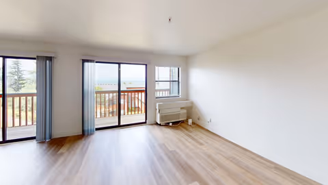 Empty room with light wood flooring, white walls, and large sliding glass doors leading to a balcony with a view of trees and distant water. A window with blinds and a wall-mounted heater are visible on the right side.