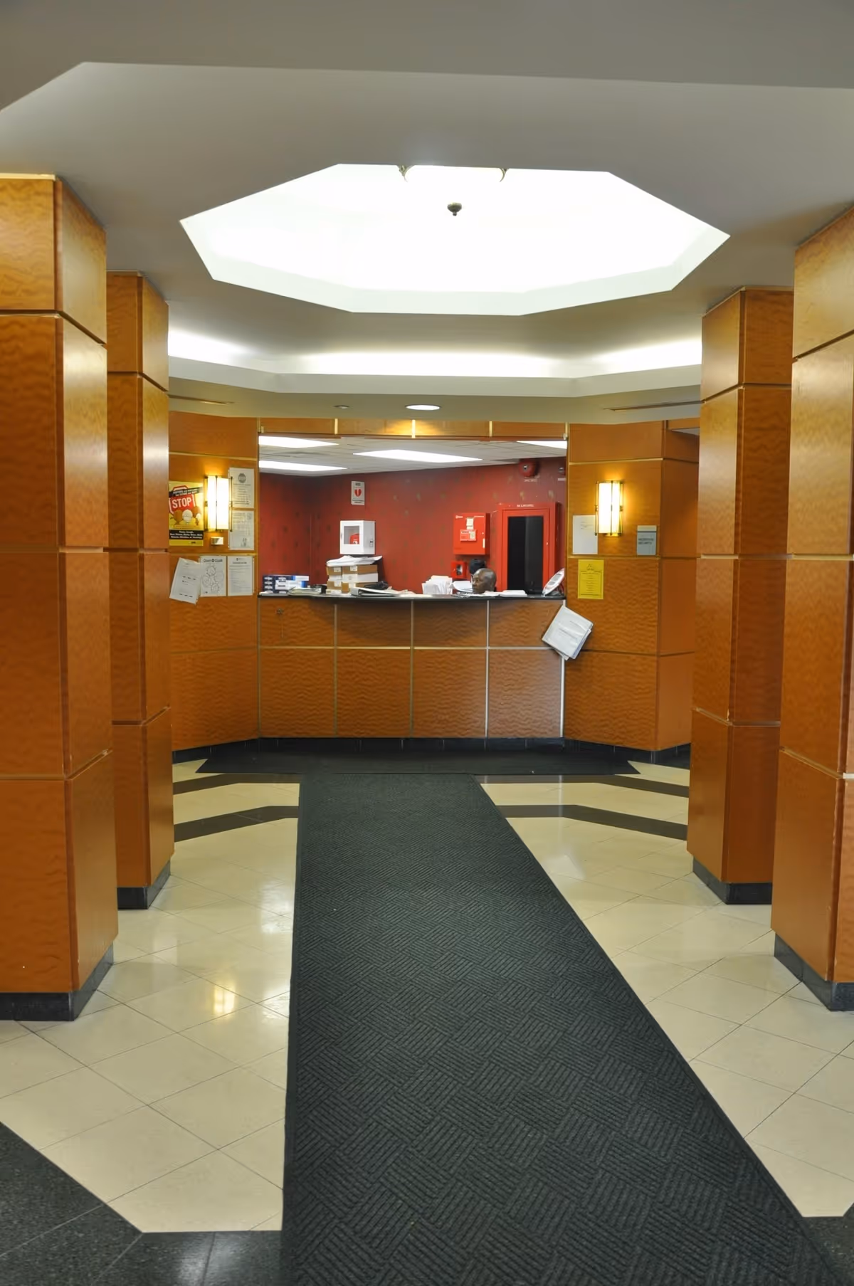 Interior view of a reception area with a front desk, wood-paneled walls, and a large octagonal ceiling light. The floor has a patterned carpet runner leading to the desk, which has papers and office supplies on it.