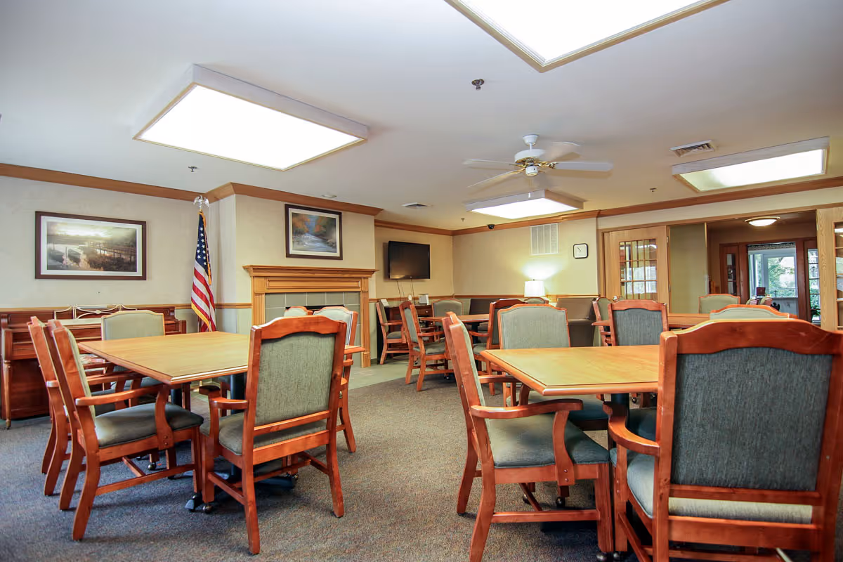 A well-lit common room with multiple wooden tables and cushioned chairs arranged neatly. The room features a fireplace with framed artwork above it, an American flag in the corner, a wall-mounted television, and ceiling fans. The walls are decorated with additional framed pictures, and there is a clock on one wall. The room has a carpeted floor and a cozy, inviting atmosphere.