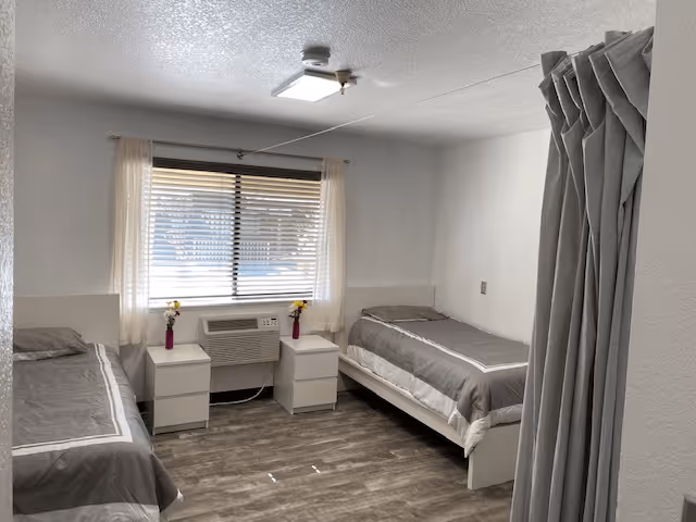 A simple, clean bedroom with two single beds on either side of a window. Each bed has gray bedding with white trim. Between the beds are two white nightstands with small vases holding yellow flowers. The window has white curtains and blinds. The floor is wood-patterned, and a gray privacy curtain is partially visible on the right side of the image.