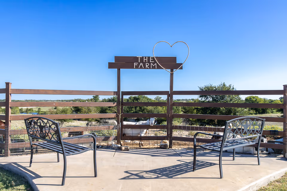 Two black metal benches facing each other on a concrete patio area with a wooden fence behind them. A sign on the fence reads 'THE FARM' with a heart shape above it. Behind the fence, there is a goat and a scenic view of trees and open land under a clear blue sky.