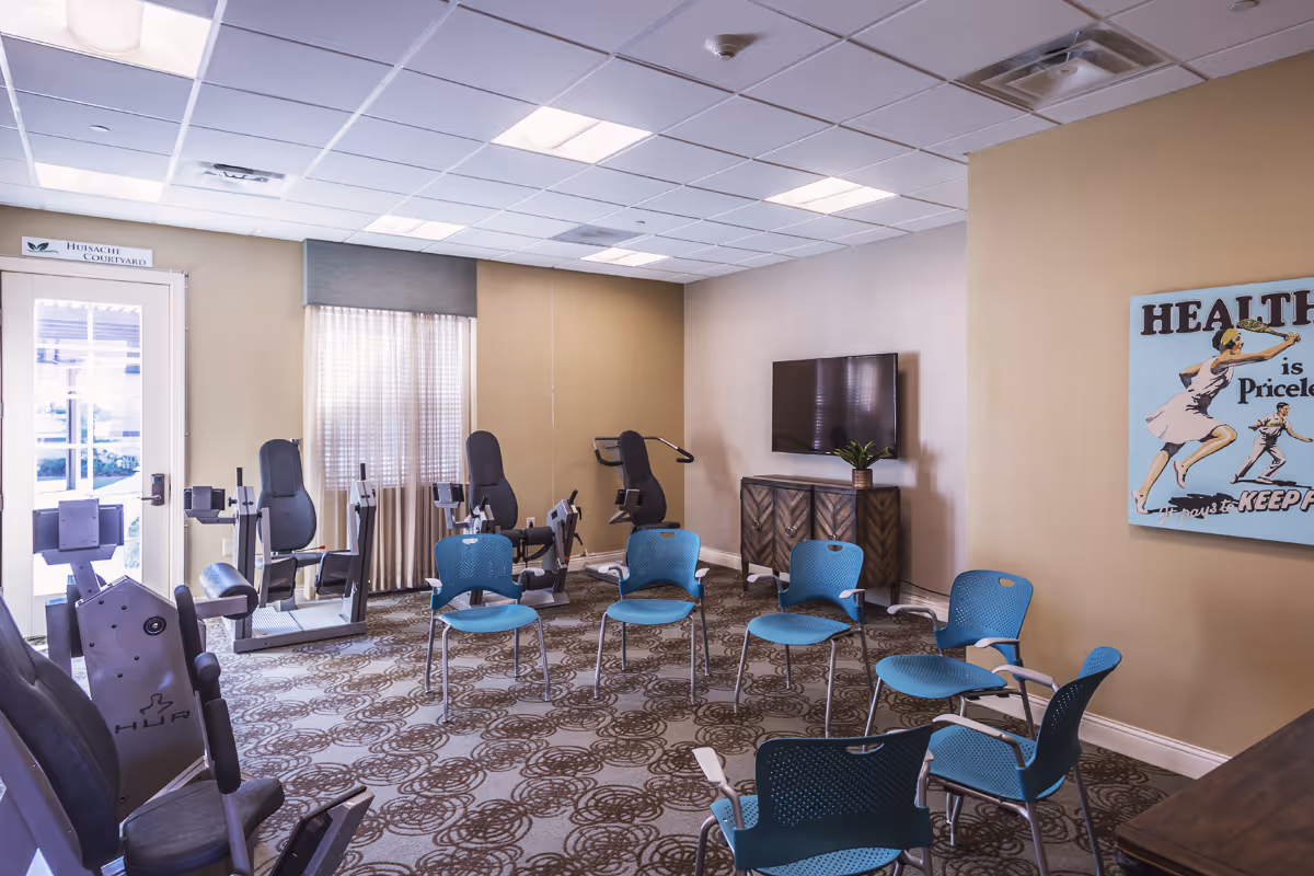 A fitness room with exercise equipment including stationary bikes and chairs arranged in a circle. There is a TV mounted on the wall above a wooden cabinet, a window with curtains, and a door labeled 'Hibachi Courtyard'. A vintage-style poster on the wall reads 'Health is Priceless'.