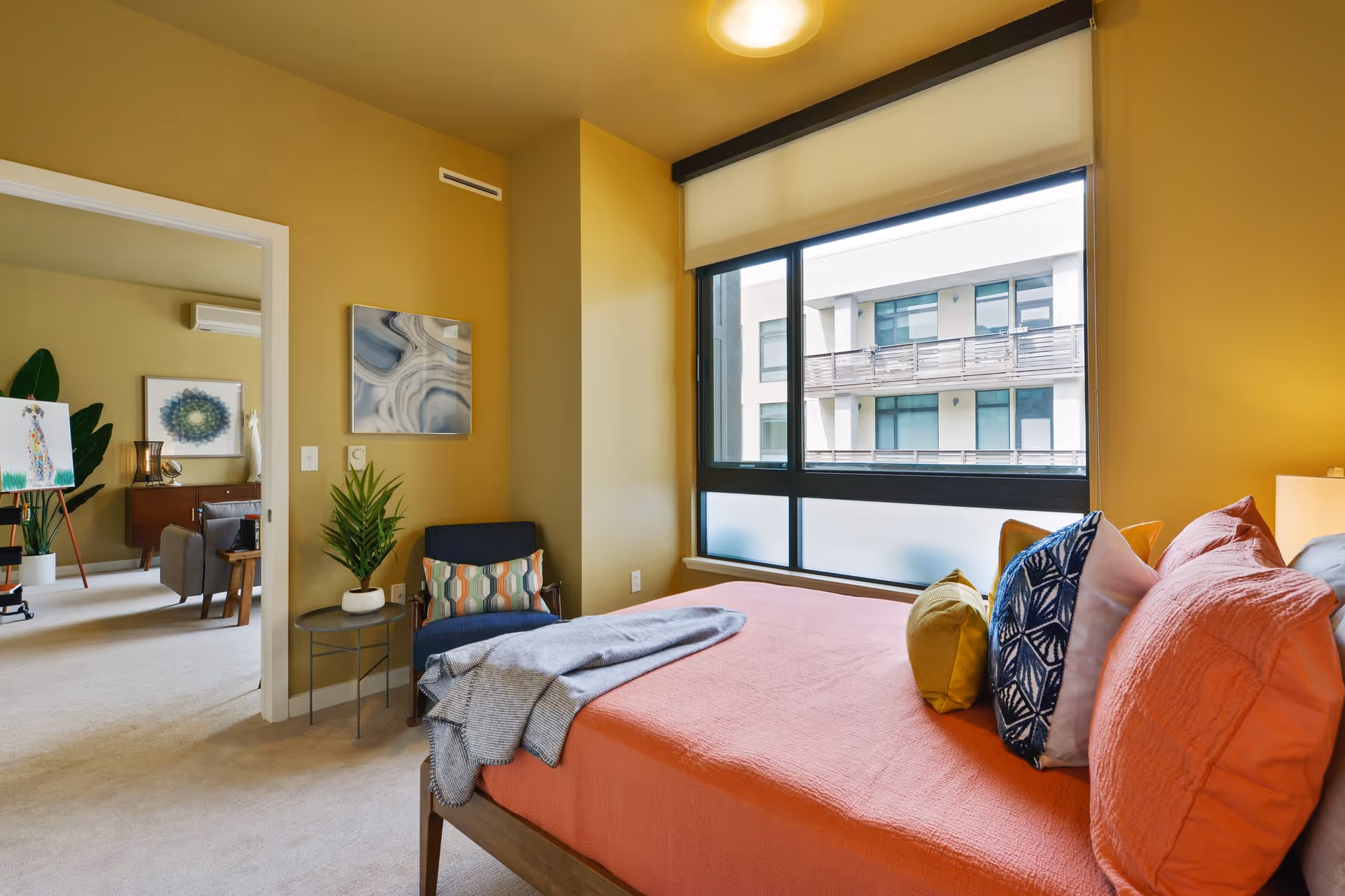 A cozy bedroom with a large window showing an exterior building. The bed has an orange bedspread with multiple colorful pillows. There is a blue armchair with a patterned cushion next to a small round table holding a potted plant. The walls are painted a warm yellow color. Through an open doorway, a living area with artwork, a plant, and furniture is visible.