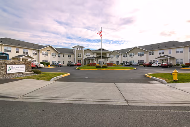 Front exterior view of Brookdale College Place, a two-story senior living facility with a central entrance, an American flag on a flagpole, parked cars, and a sign displaying the facility's name on the left side.