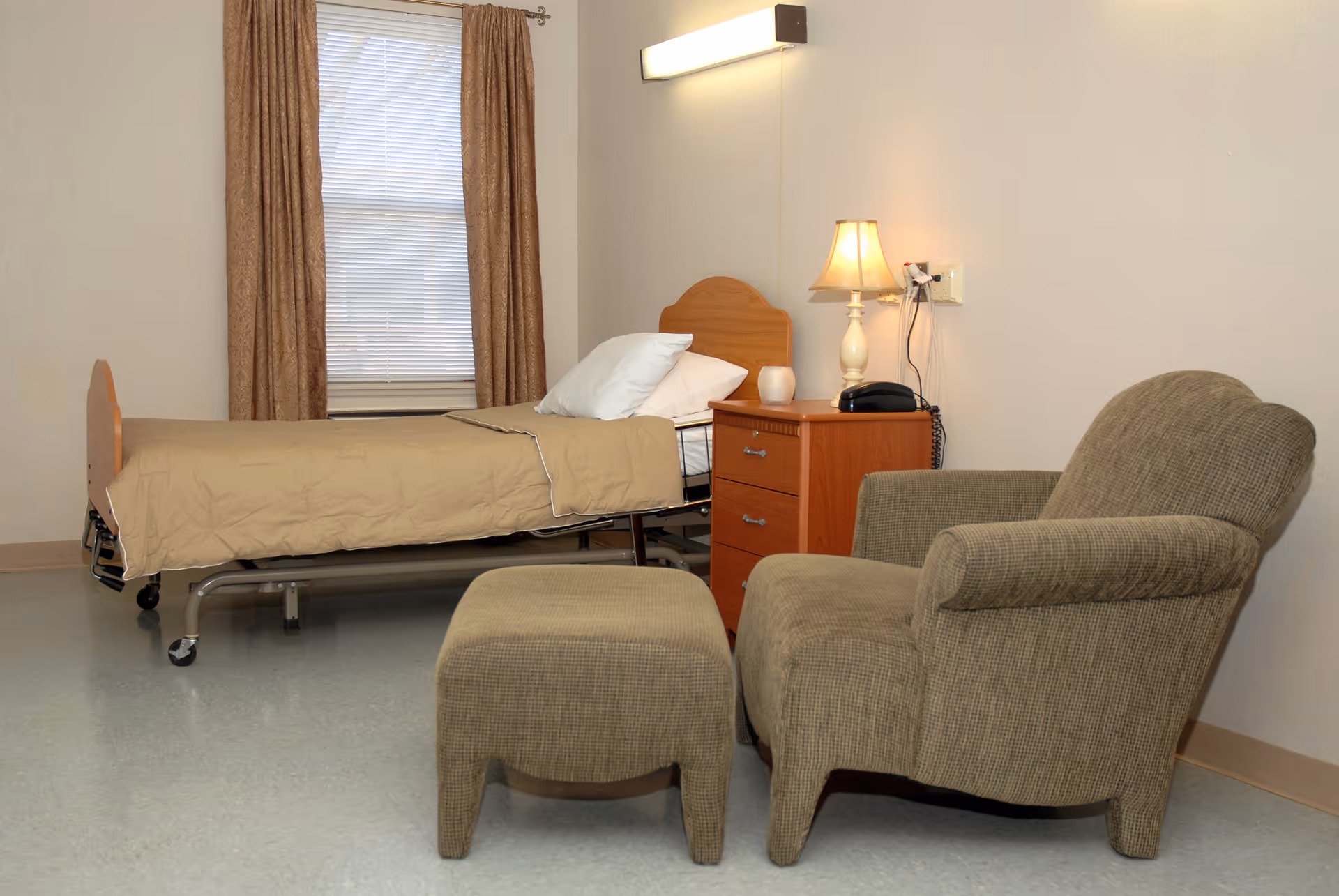 A simple room with a single hospital-style bed covered with a beige blanket and white pillow, a wooden nightstand with a lamp and telephone, and a cushioned armchair with a matching ottoman. A window with closed blinds and brown curtains is visible behind the bed.