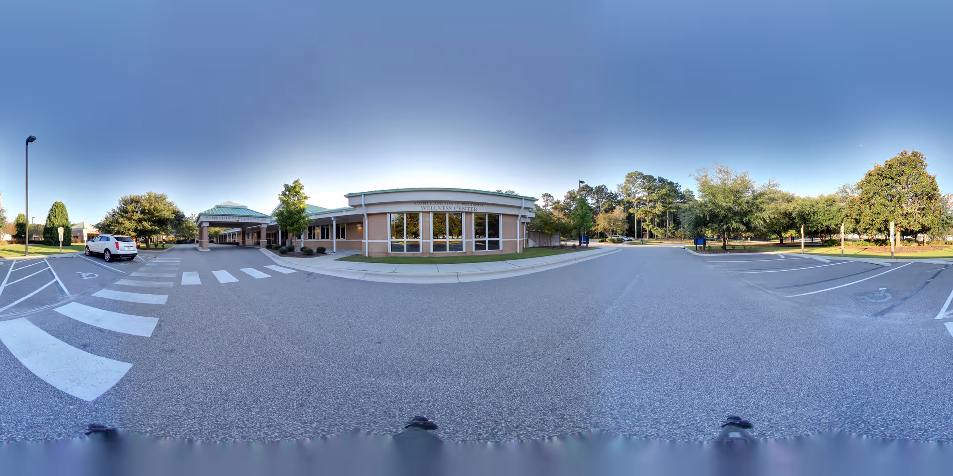 Wide panoramic view of the exterior front entrance of The Davis Community facility, showing a single-story brick building with large windows, a covered entryway, a parking lot with marked spaces including handicapped spots, and surrounding trees and greenery under a clear blue sky.