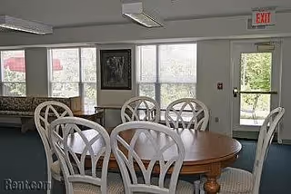 Interior view of a senior living facility dining area with a round wooden table surrounded by eight white chairs with curved backs. Large windows and a glass door provide natural light and a view of greenery outside. A framed picture hangs on the wall, and an exit sign is visible above the door.