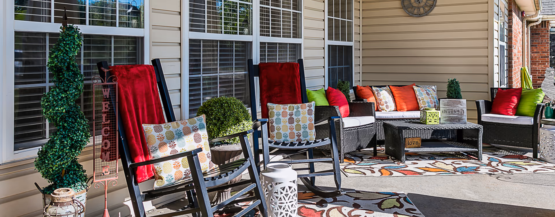 Outdoor patio area with black rocking chairs and wicker seating adorned with colorful cushions and red throws. Decorative plants, a welcome sign, and a clock on the beige siding wall are also visible, along with patterned outdoor rugs on the concrete floor.