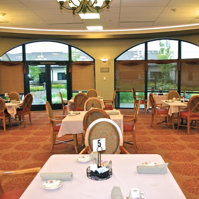 A dining room in an assisted living facility with multiple tables covered in light pink tablecloths, each set with plates, cups, napkins, and utensils. The room has large arched windows with blinds partially drawn, allowing natural light to enter. Chairs with patterned upholstery surround the tables, and a table number 5 is visible in the foreground.
