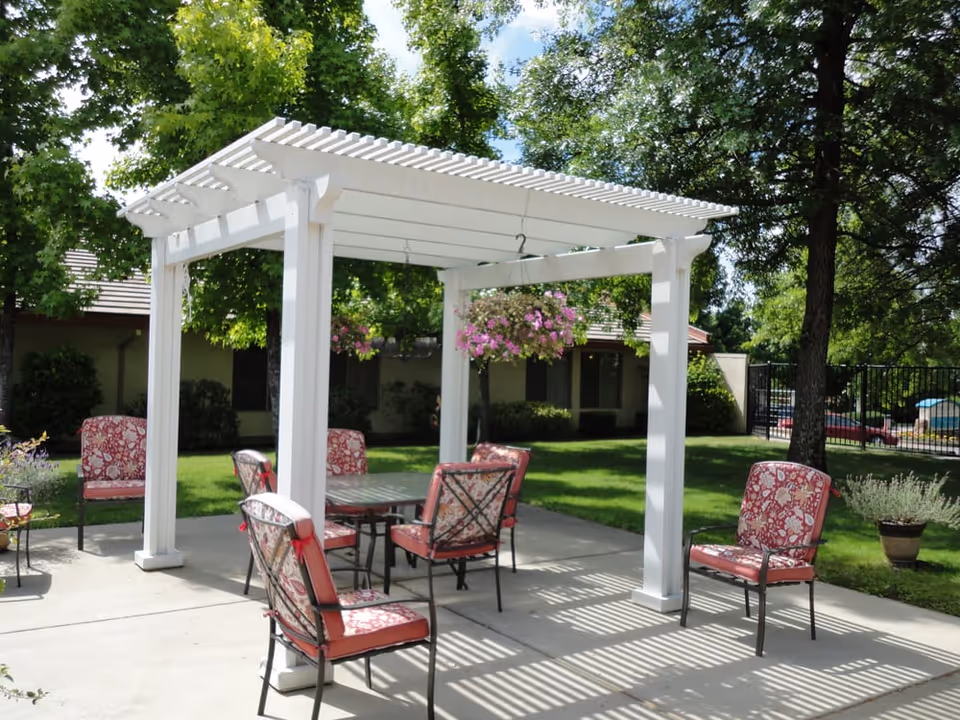 Outdoor patio area with a white pergola providing partial shade over a glass-top table surrounded by six cushioned chairs with red and white floral patterns. Hanging flower baskets with pink blooms are suspended from the pergola. The patio is surrounded by green grass, trees, and shrubs, with a building and a black metal fence visible in the background under a partly cloudy sky.