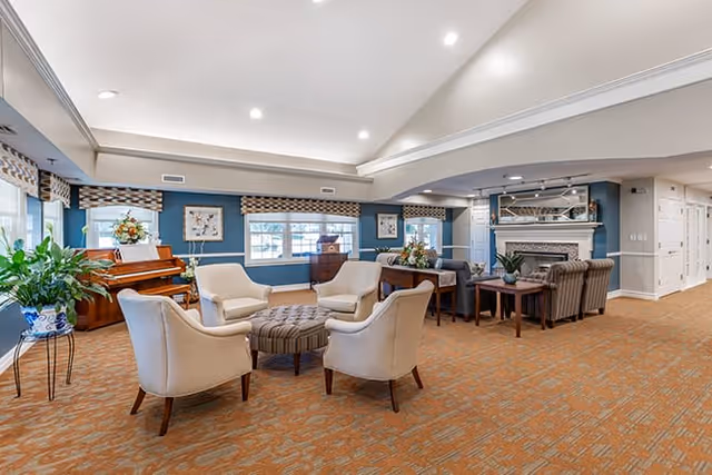 A spacious and well-lit living room area in an assisted living facility featuring a group of four beige armchairs arranged around a round ottoman. The room has blue walls with windows adorned with patterned valances, a wooden piano with a floral arrangement on top, framed artwork, and a fireplace with a mantel decorated with plants and other items. The floor is covered with a patterned carpet, and there are additional seating areas with sofas and tables.