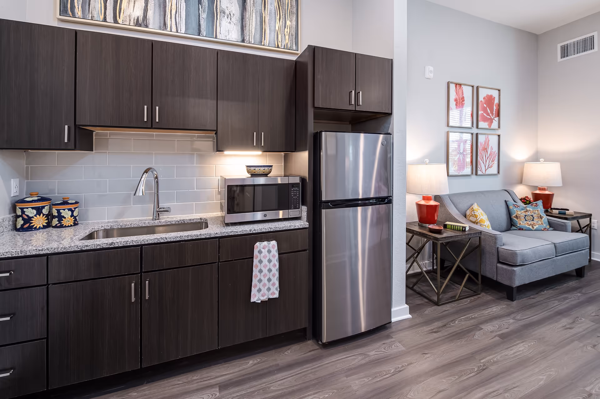 Modern kitchen area with dark wood cabinets, a granite countertop, a stainless steel refrigerator, a microwave, and a sink. Adjacent to the kitchen is a small living area with a gray loveseat, two side tables with red lamps, and four framed red and pink botanical prints on the wall.