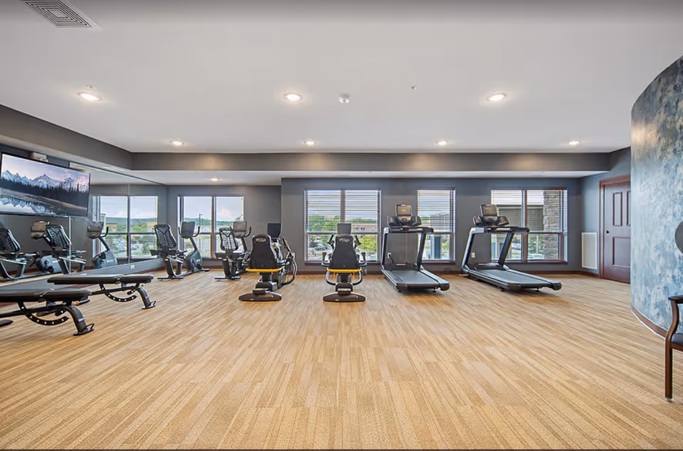 Spacious exercise room with treadmills, recumbent bikes, and benches facing large windows.
