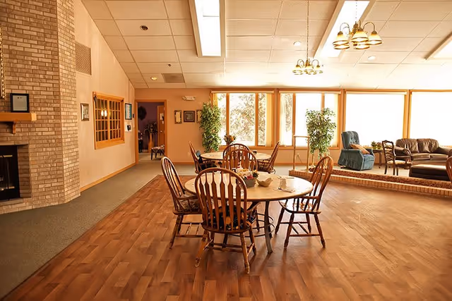 Sunlit common room with a round wooden dining table and chairs, lounge seating, and large windows.