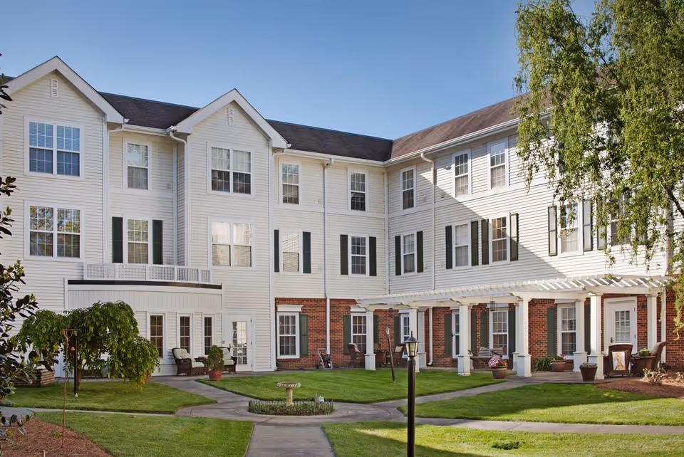 Exterior view of a three-story senior living facility building with white siding and brick accents. The building surrounds a well-maintained courtyard with green grass, a few small trees, potted plants, and a paved walkway. There are several windows with green shutters and a pergola-covered patio area with chairs.
