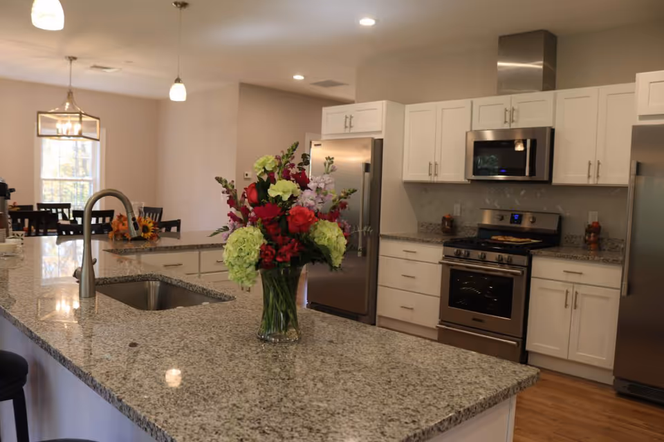 Modern kitchen with a large granite island, sink, stainless steel appliances, white cabinets, and a vase of flowers on the counter.