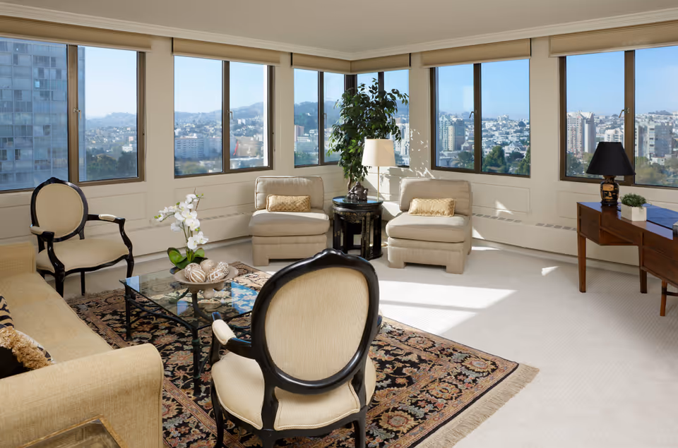 A bright living room with large windows offering a panoramic city view. The room features beige upholstered chairs and a sofa, a glass coffee table with decorative items, a patterned area rug, a wooden side table with a black lamp, and a tall green plant near the corner windows.