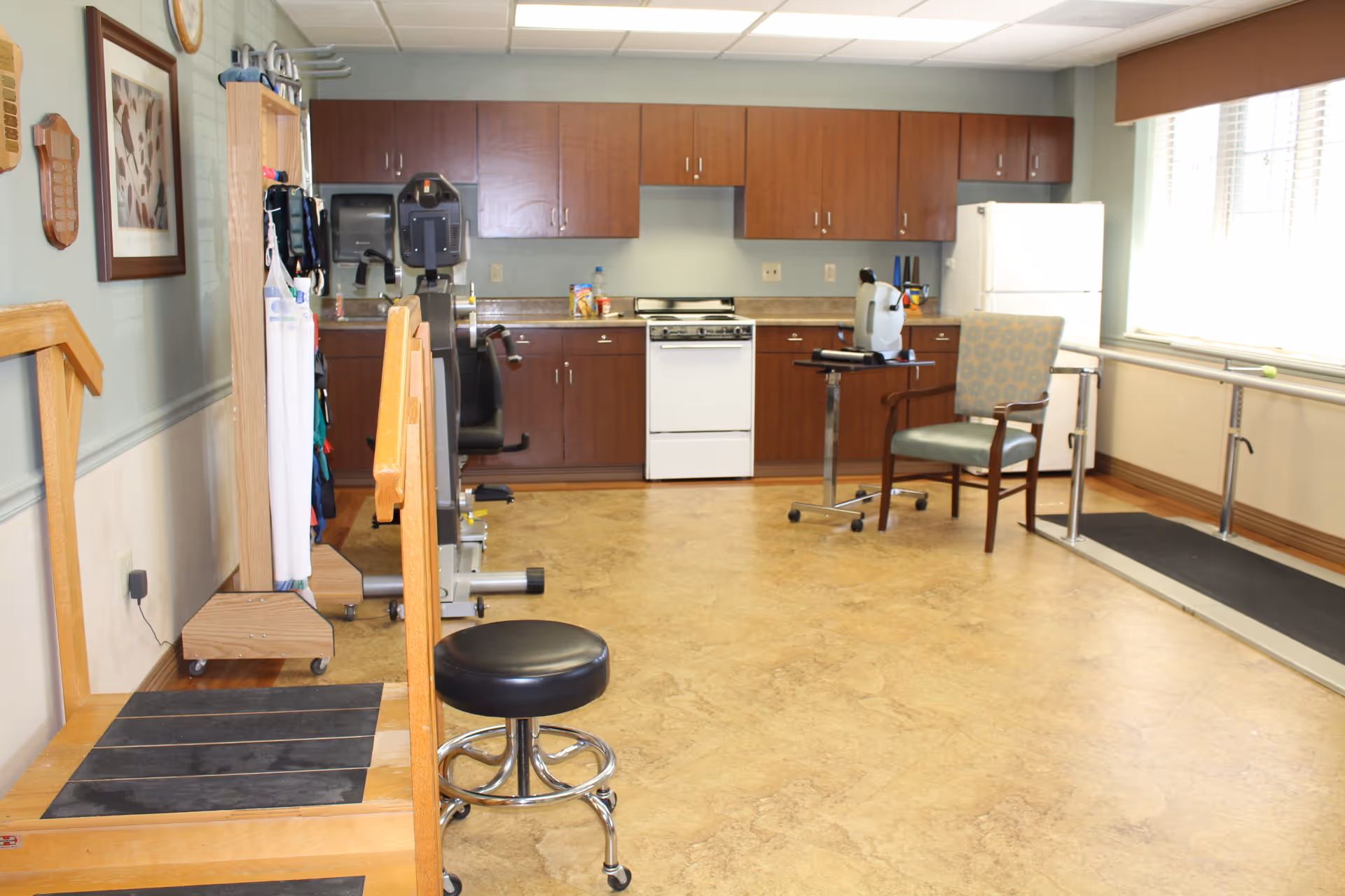 A rehabilitation or therapy room with exercise equipment, a wooden step platform, a stool, a chair, parallel bars for walking practice, and a kitchenette with wooden cabinets, a stove, and a refrigerator.