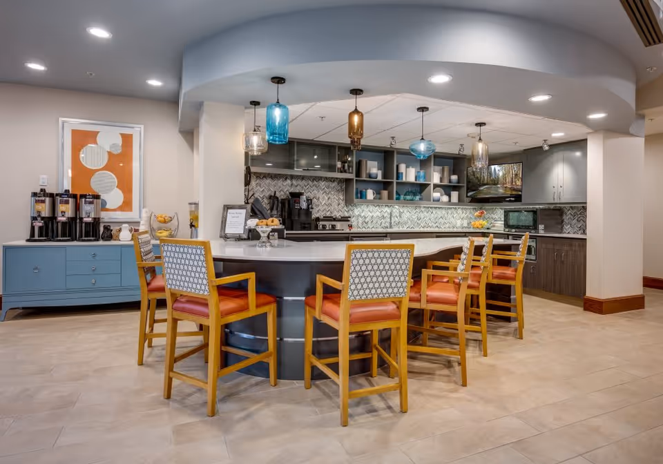 A modern kitchen area with a curved island countertop surrounded by six wooden chairs with orange cushions and patterned backs. The kitchen features a tiled backsplash, open shelves with decorative items, a coffee station on a blue sideboard, and pendant lights hanging from the ceiling. The floor is tiled and the space is well-lit with recessed lighting.