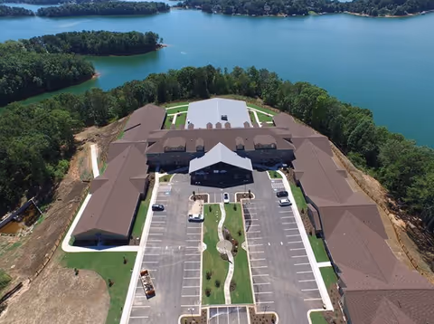 Aerial view of The Phoenix at Lake Lanier senior living facility showing a large building complex with multiple wings surrounding a central parking lot. The facility is situated near a large body of water with trees and greenery surrounding the property.