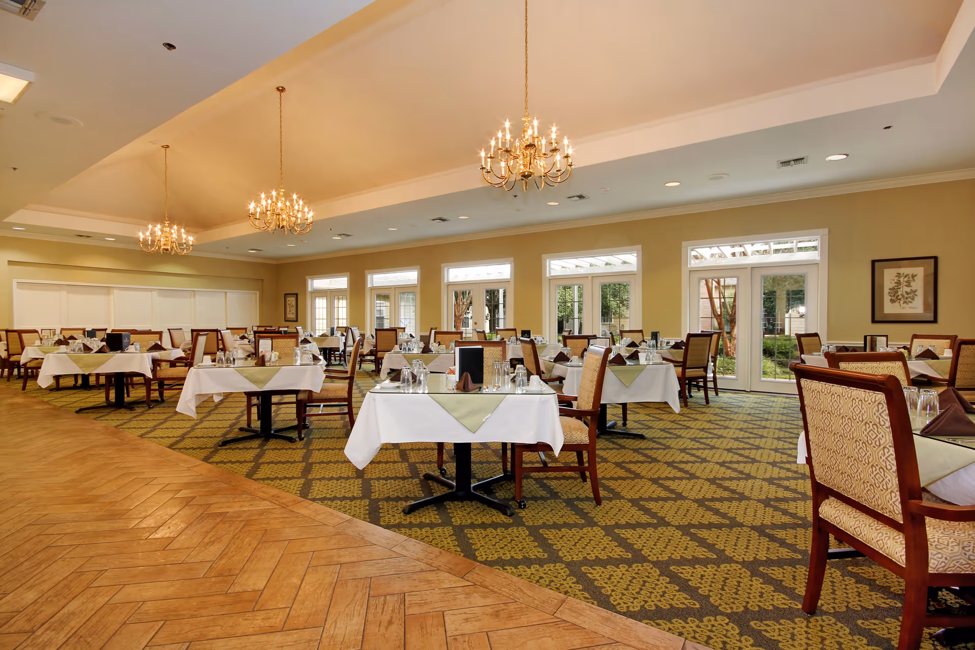 Spacious, well-lit dining room with multiple set tables and chairs, chandeliers, and large windows looking out to a courtyard.