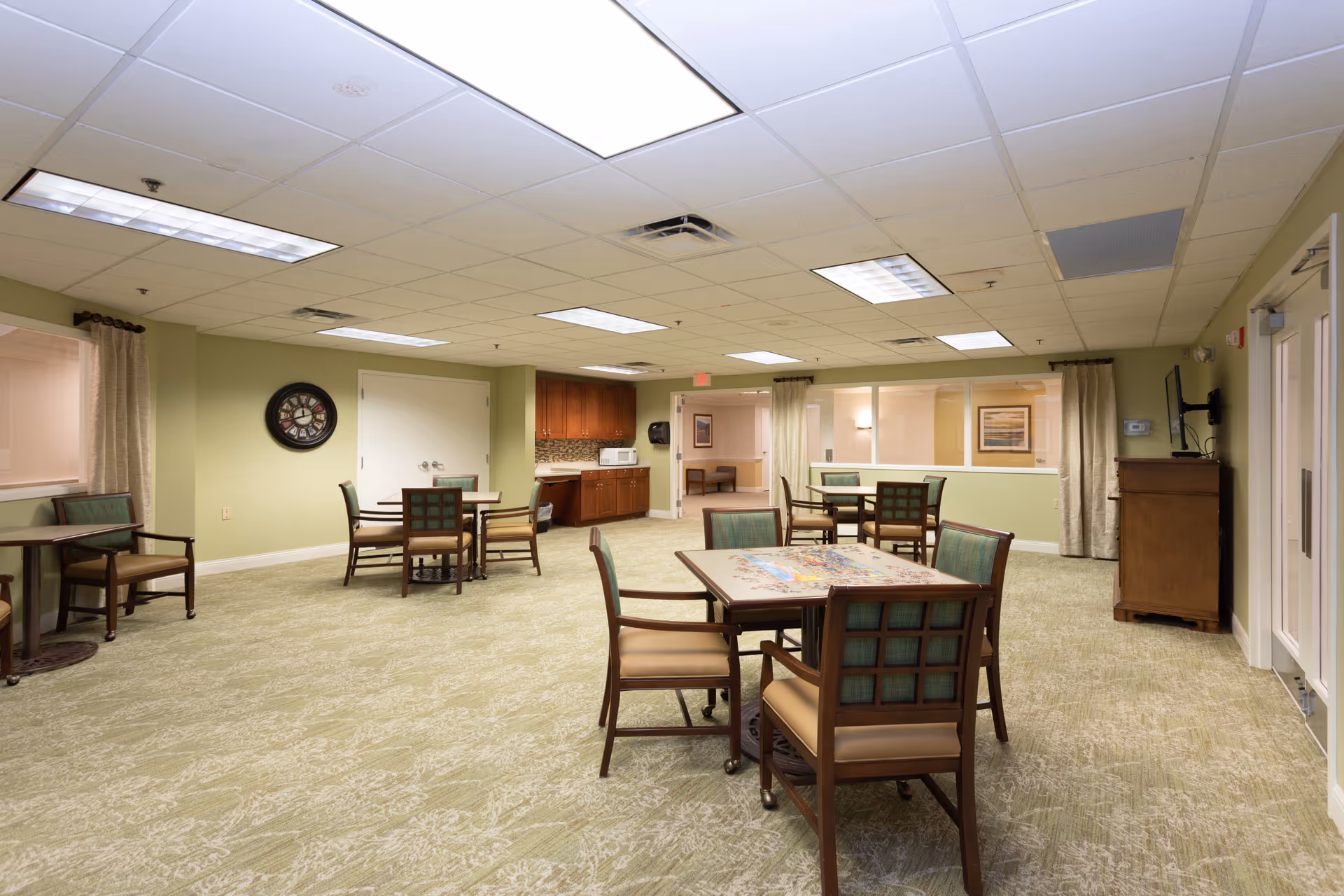 A spacious common room with multiple tables and chairs arranged for group activities or dining. The room has light green walls, a patterned carpet, and a drop ceiling with fluorescent lighting. There is a small kitchenette area with wooden cabinets, a microwave, and a sink. A clock is mounted on one wall, and a television is placed on a wooden stand near the entrance door. Windows with curtains and framed artwork are visible on the walls.