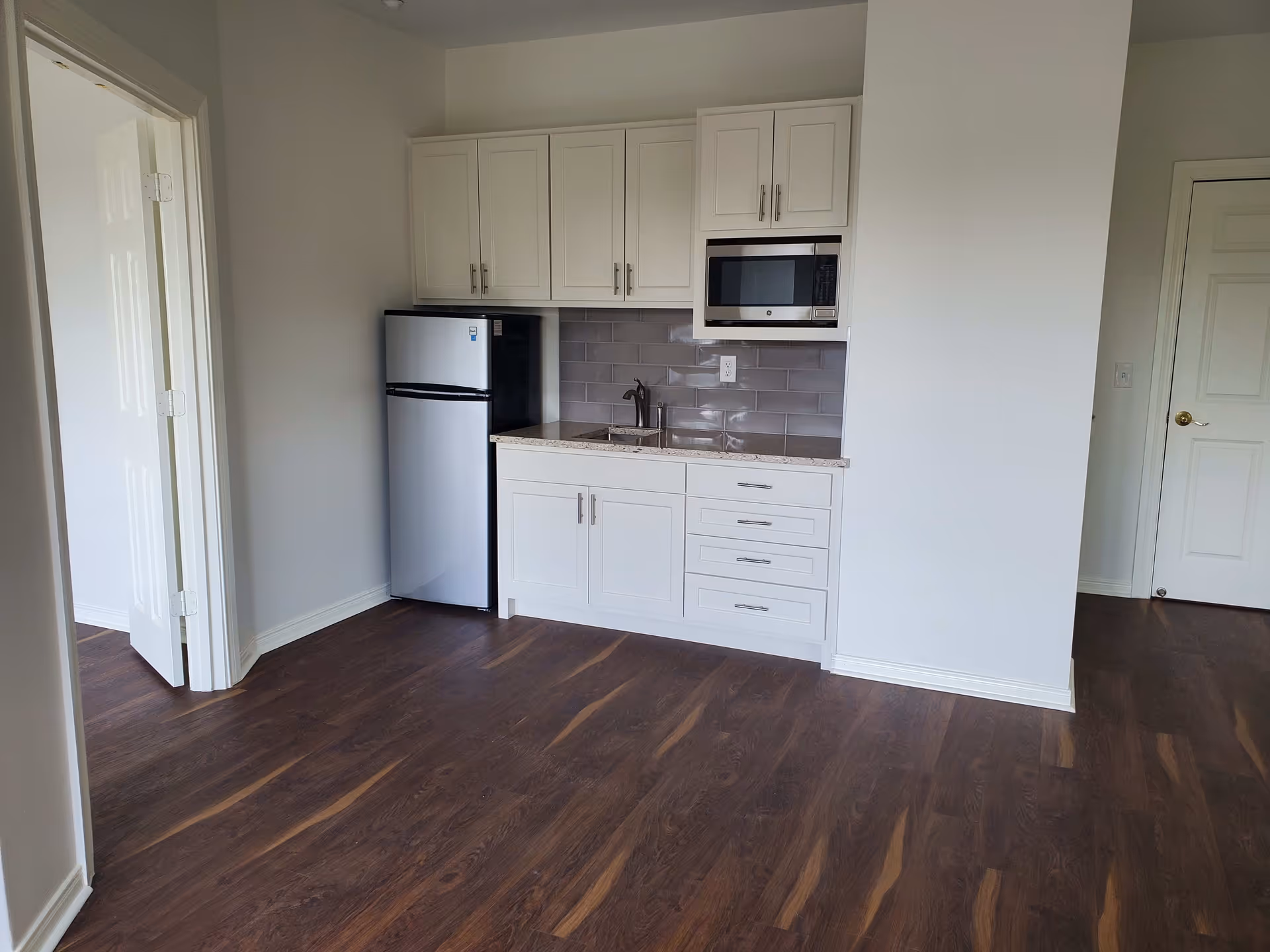 Compact kitchenette with white cabinets, a stainless-steel mini refrigerator and microwave above a sink, gray subway-tile backsplash and dark wood flooring.
