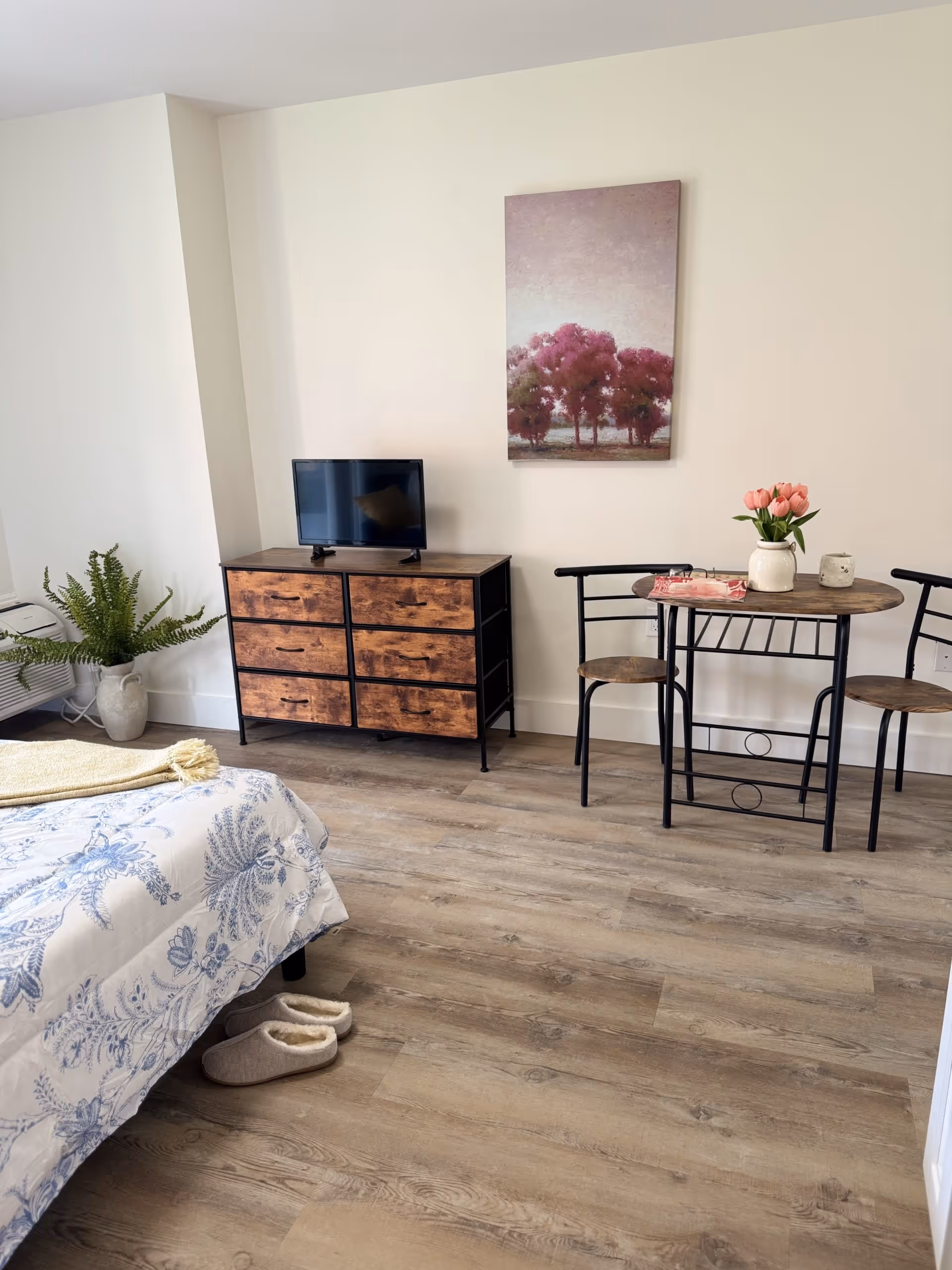 A cozy bedroom corner featuring a bed with a blue and white floral comforter, a pair of slippers on the wooden floor, a wooden dresser with a small TV on top, a small round table with two chairs, a vase with pink flowers, and a painting of pink trees on the wall.