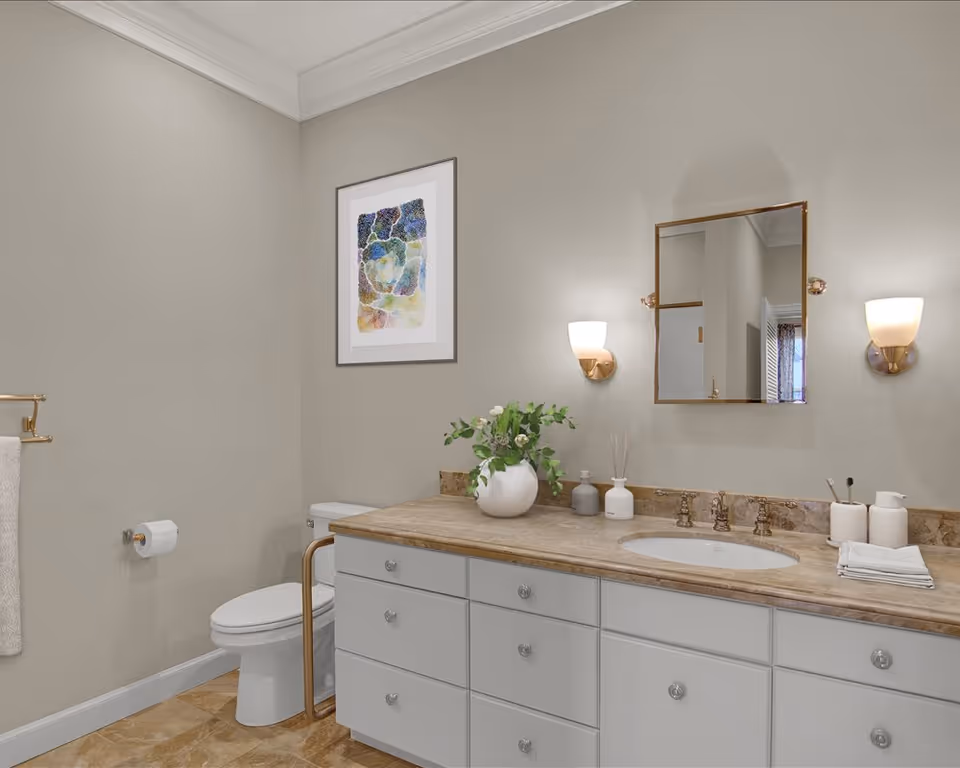 A clean and modern bathroom featuring a white toilet, a beige marble countertop with an under-mount sink, and a large vanity with multiple drawers. Above the sink is a rectangular mirror flanked by two wall-mounted light fixtures. A white vase with green foliage and other small decorative items are placed on the countertop. A towel rack with a white towel and a toilet paper holder are mounted on the beige wall. A framed abstract artwork hangs above the toilet.