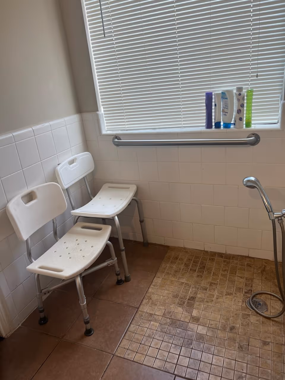 A bathroom shower area with two white plastic shower chairs with metal legs, a tiled floor with a drain, a handheld showerhead, white tiled walls, a horizontal grab bar, and a window with closed blinds. Several bottles of toiletries are placed on the window sill.
