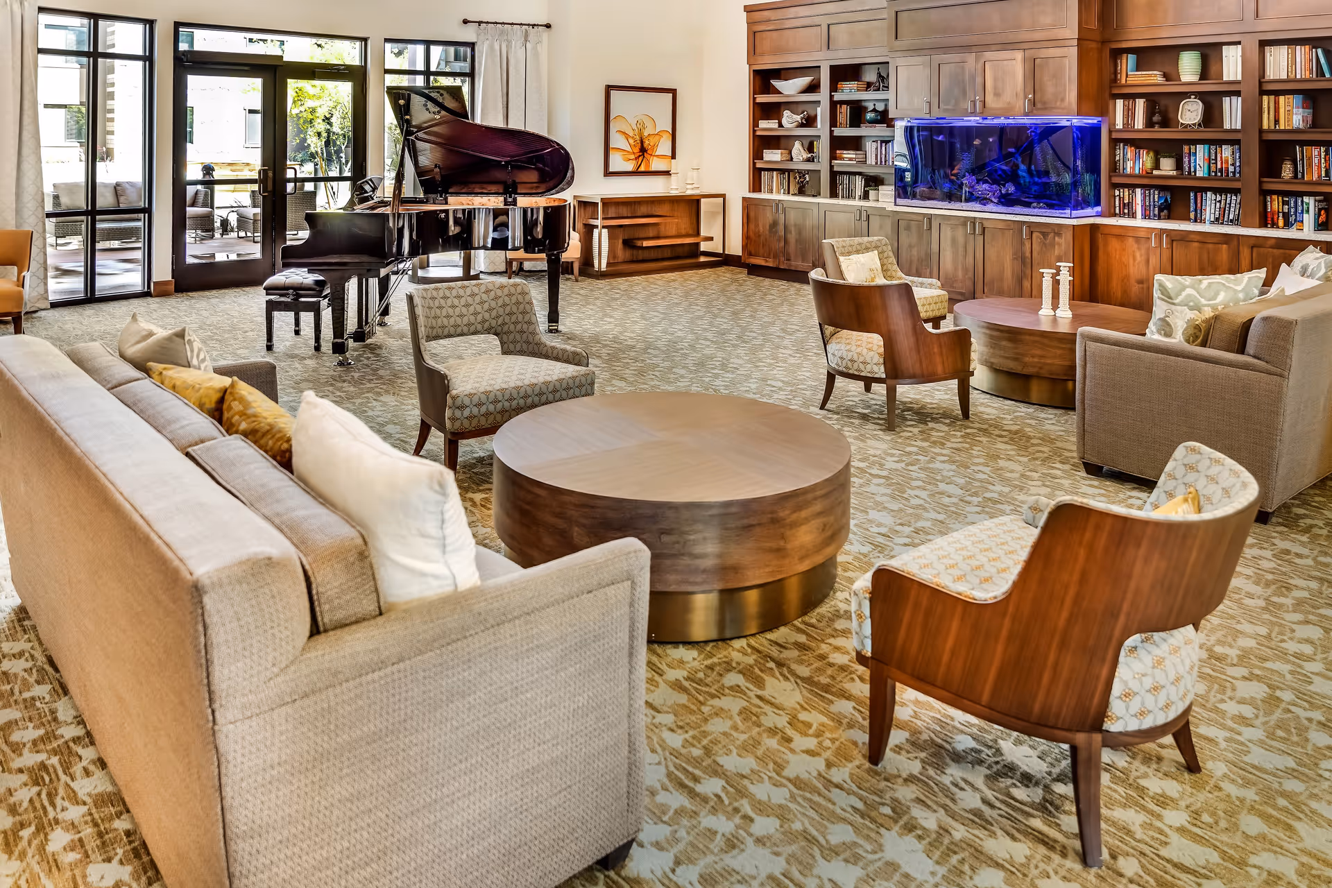 A spacious and well-lit living room area with beige sofas and patterned armchairs arranged around two round wooden coffee tables. A grand piano is positioned near large glass doors that lead to an outdoor patio with seating. The room features built-in wooden bookshelves filled with books and decorative items, and a large aquarium with blue lighting is integrated into the shelving unit.