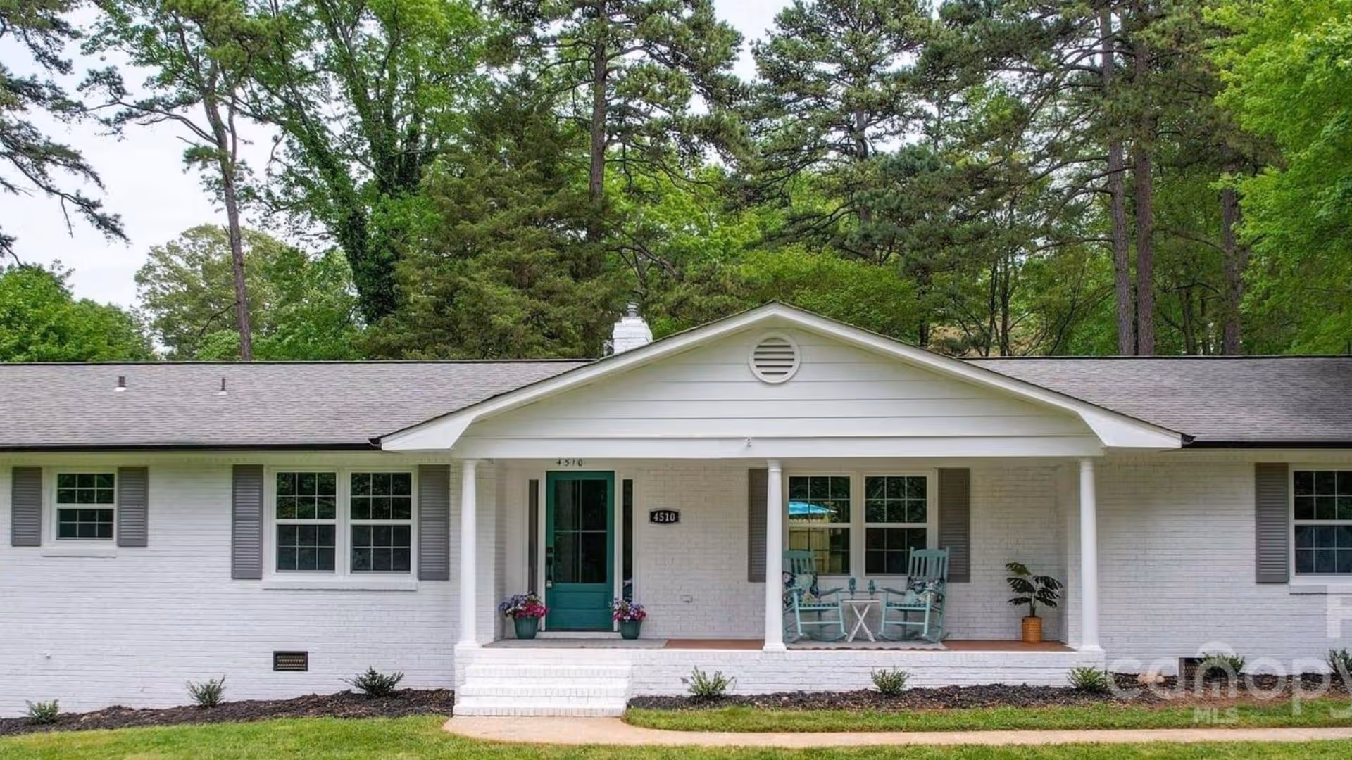 A single-story white brick building with a green front door and a covered front porch with chairs and potted plants.