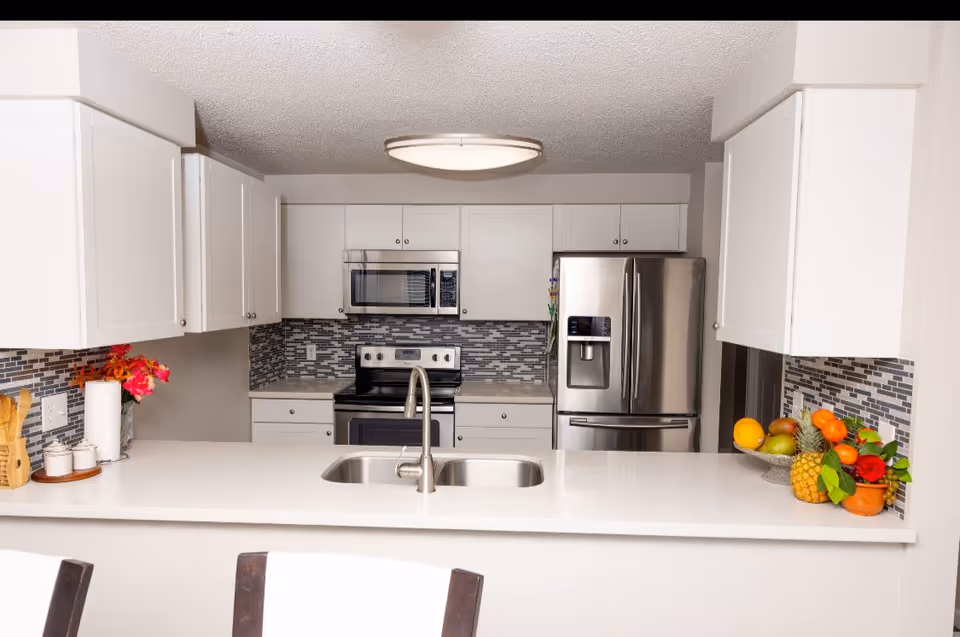 Modern kitchen with white cabinets, a stainless steel refrigerator, stove, and microwave. The countertop has a double sink with a faucet, a bowl of assorted fruits including pineapple and oranges, and a small plant. The backsplash features a mosaic tile design in shades of gray and white. There are two chairs partially visible in the foreground.